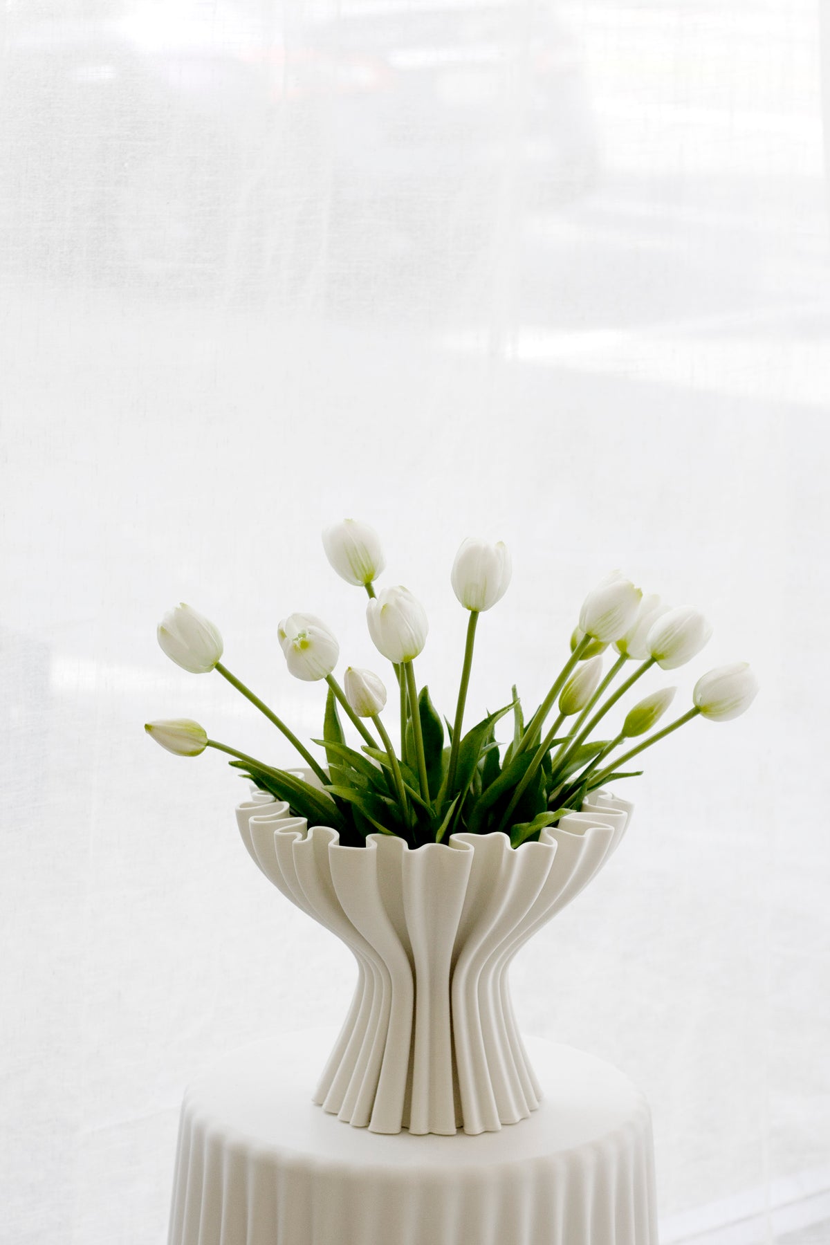 Small white faux tulips in a ribbed white vase on a fluted pedestal beside a window