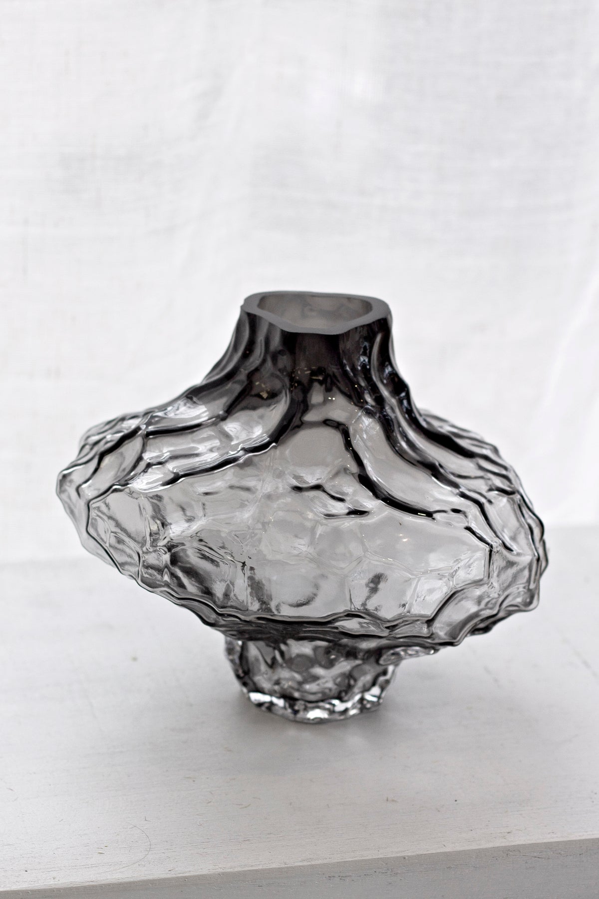 Smoky black rippled crystal glass vase on a pale bench against a linen backdrop