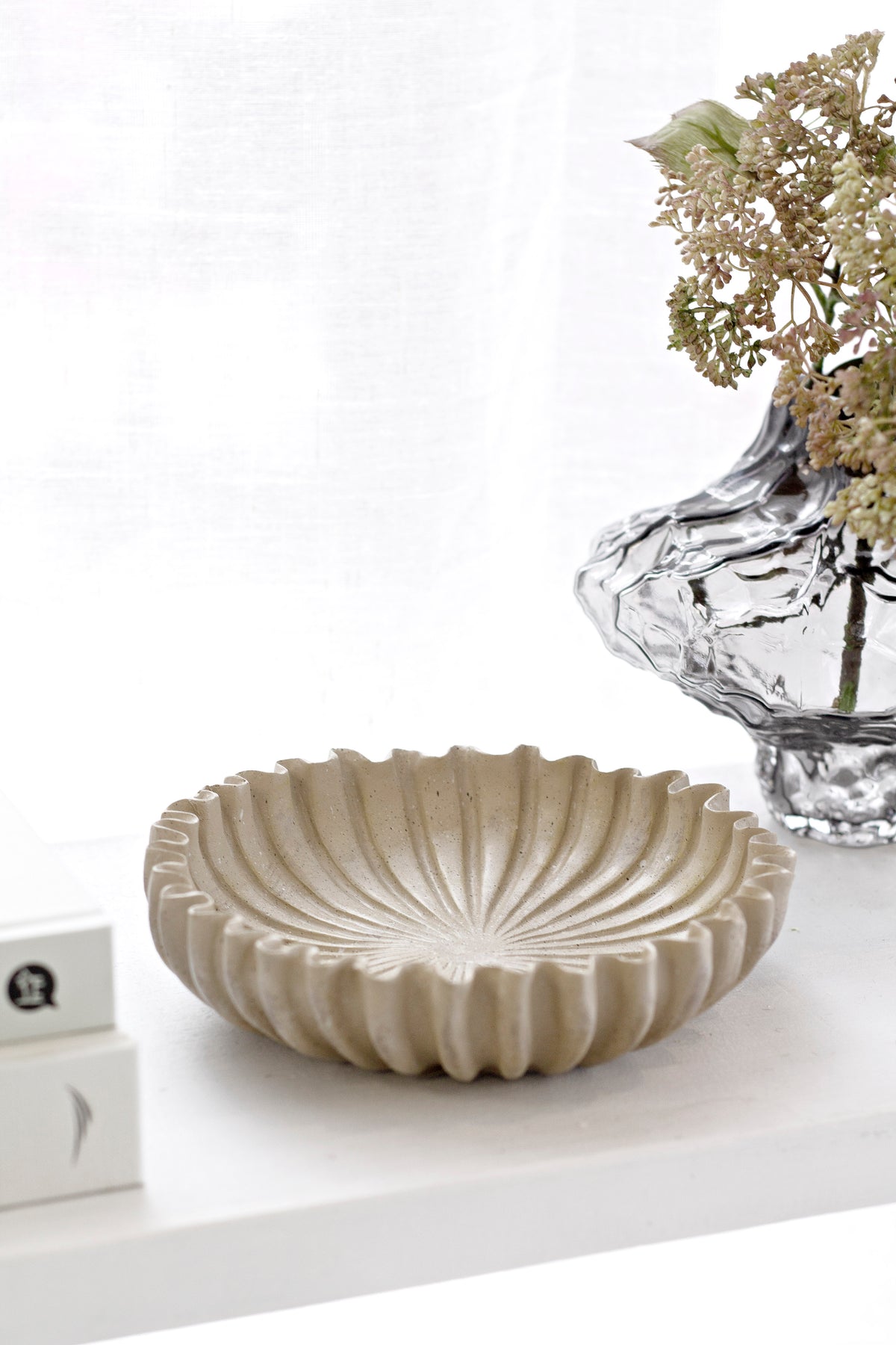 Textured scalloped-edge stoneware platter styled on an entry table beside a glass vase with dried blooms and stacked books.