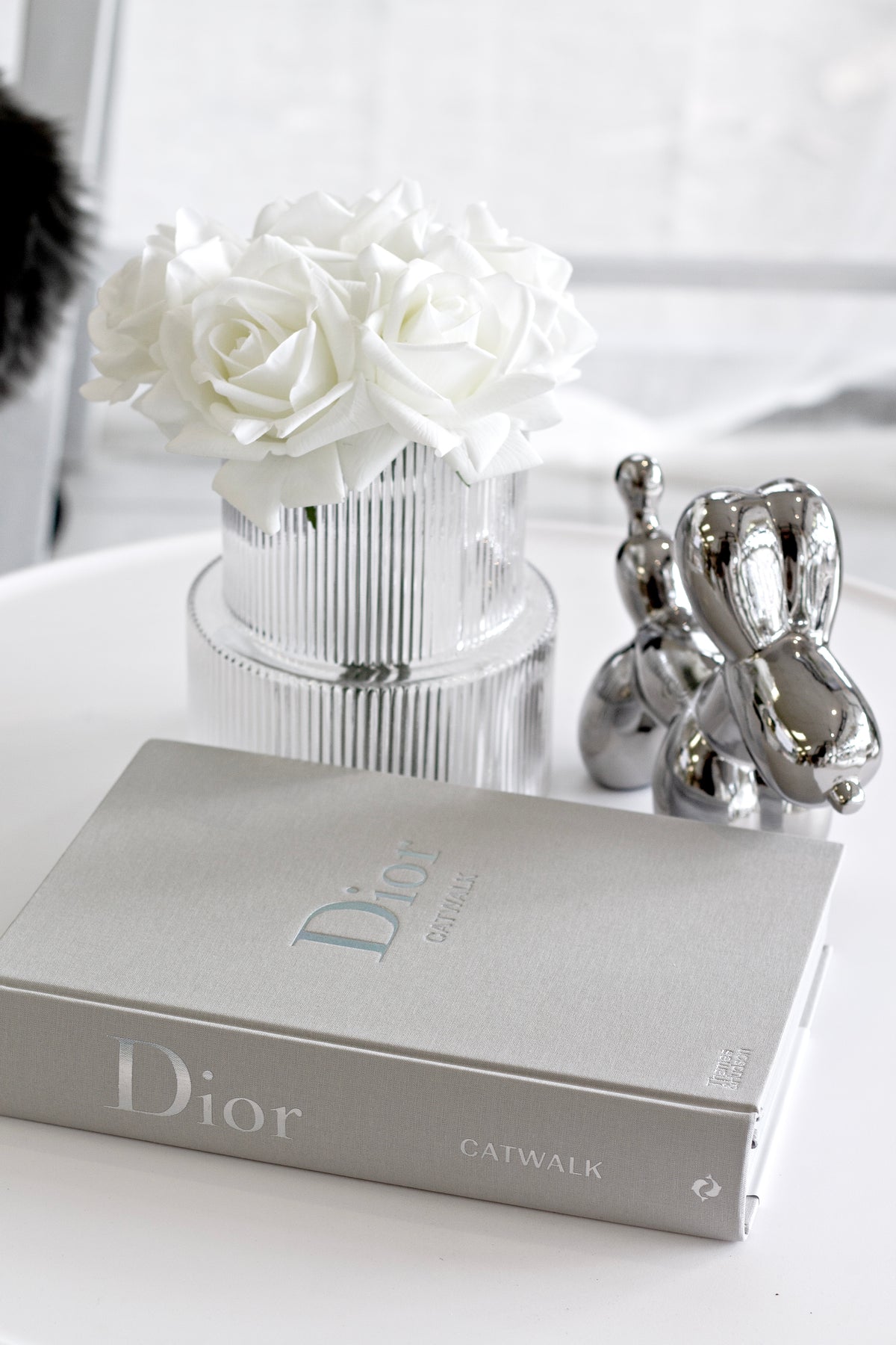 Silver ceramic balloon dog sculpture on a white side table with a Dior Catwalk book and a vase of white roses