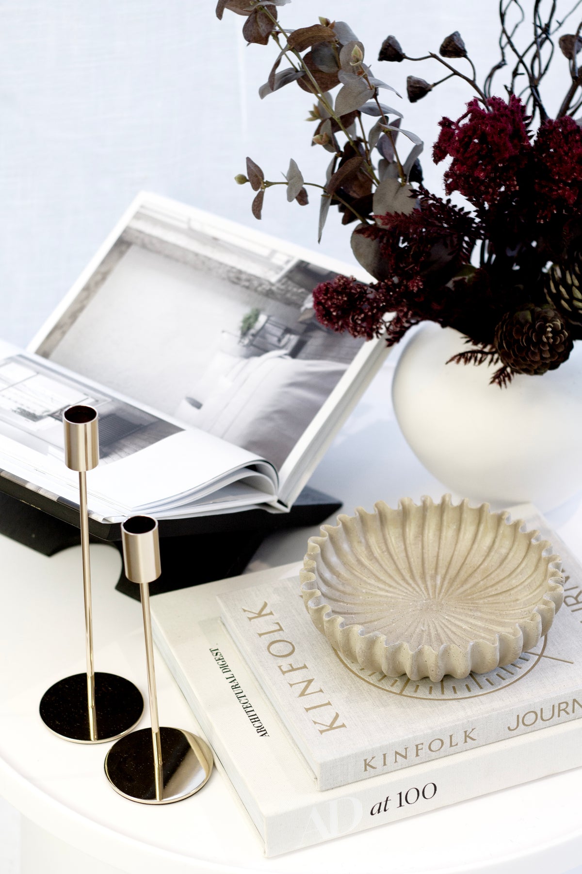 Brown eucalyptus with deep red blooms in a white vase, brass candle holders and books alongside