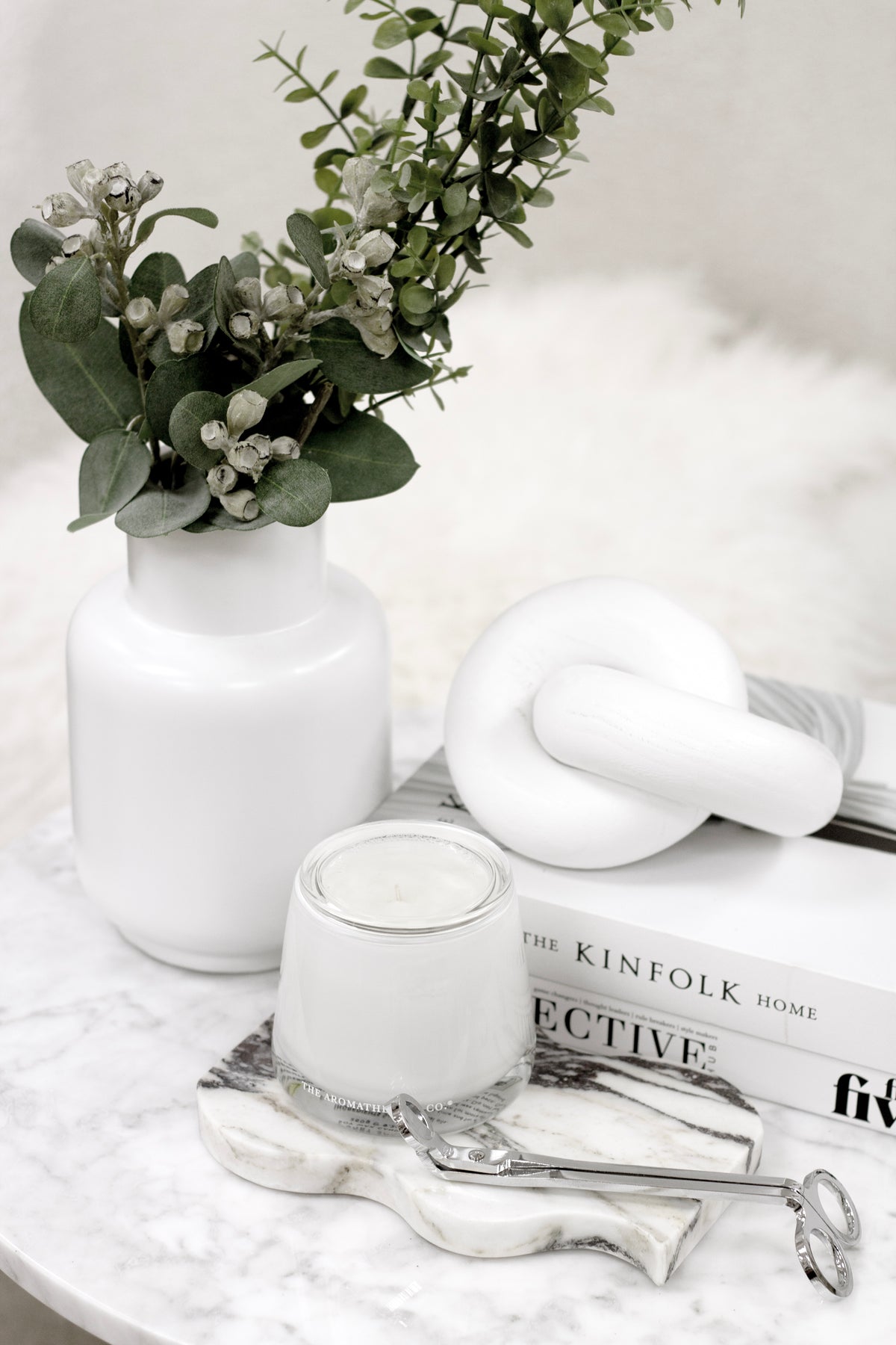 Silver wick trimmer on marble with a candle, a knot sculpture and eucalyptus in a white vase