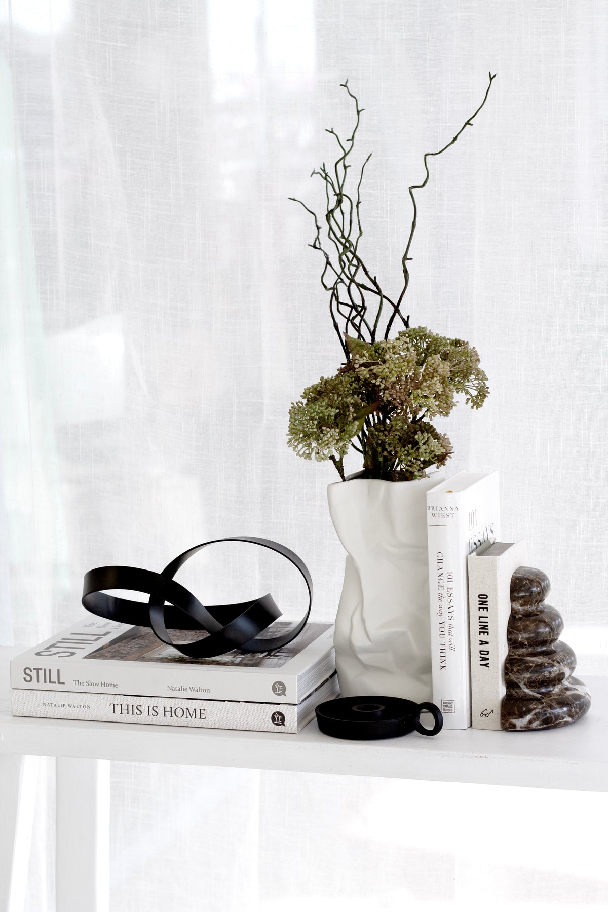 Black Infinity Loop sculpture on stacked books beside a white vase of dried hydrangea and a dark stone ornament