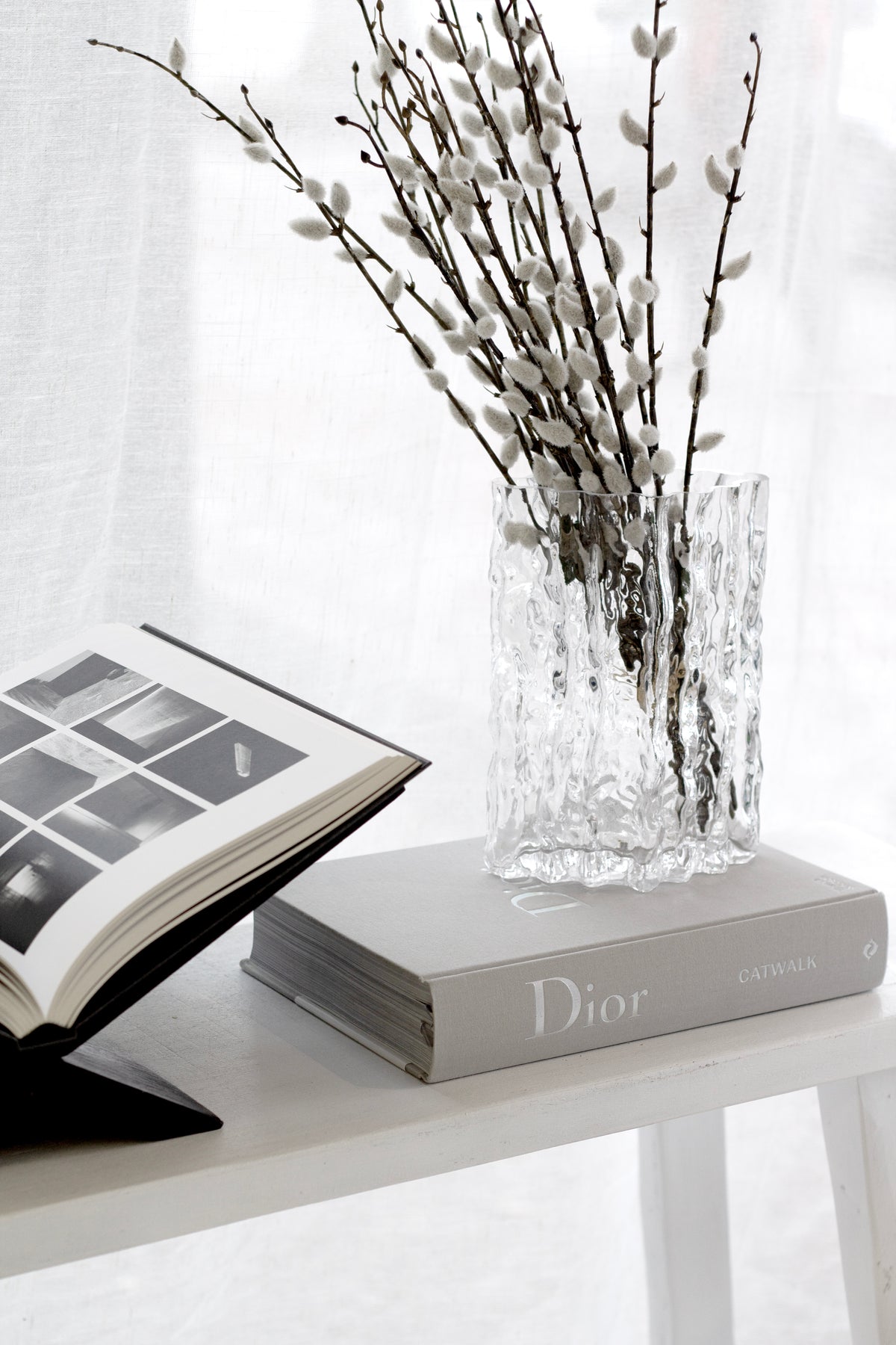 Faux pussy willow in a glass vase beside an open book on a white shelf