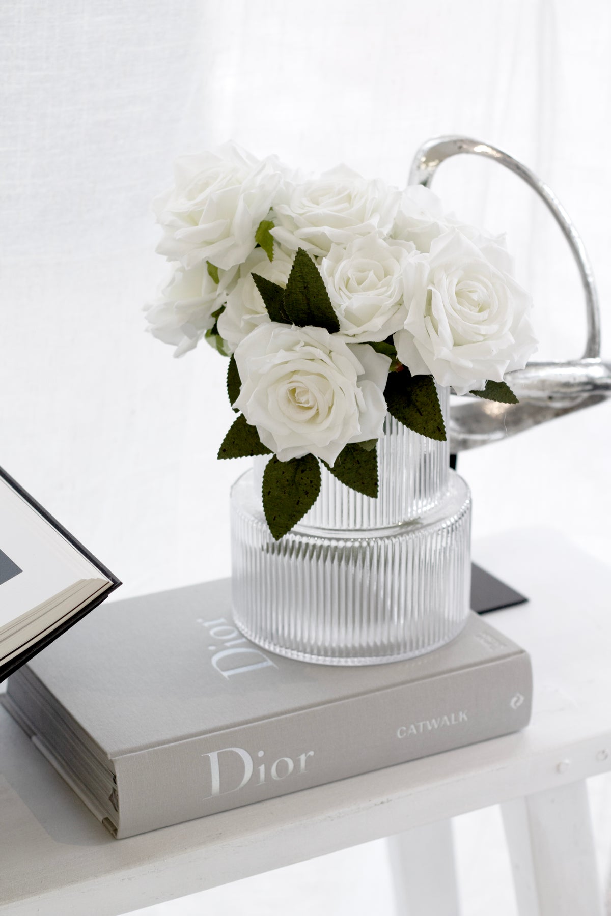 Realistic white faux rose bouquet arranged in a clear ribbed glass vase styled on a neutral book stack with minimal decor.