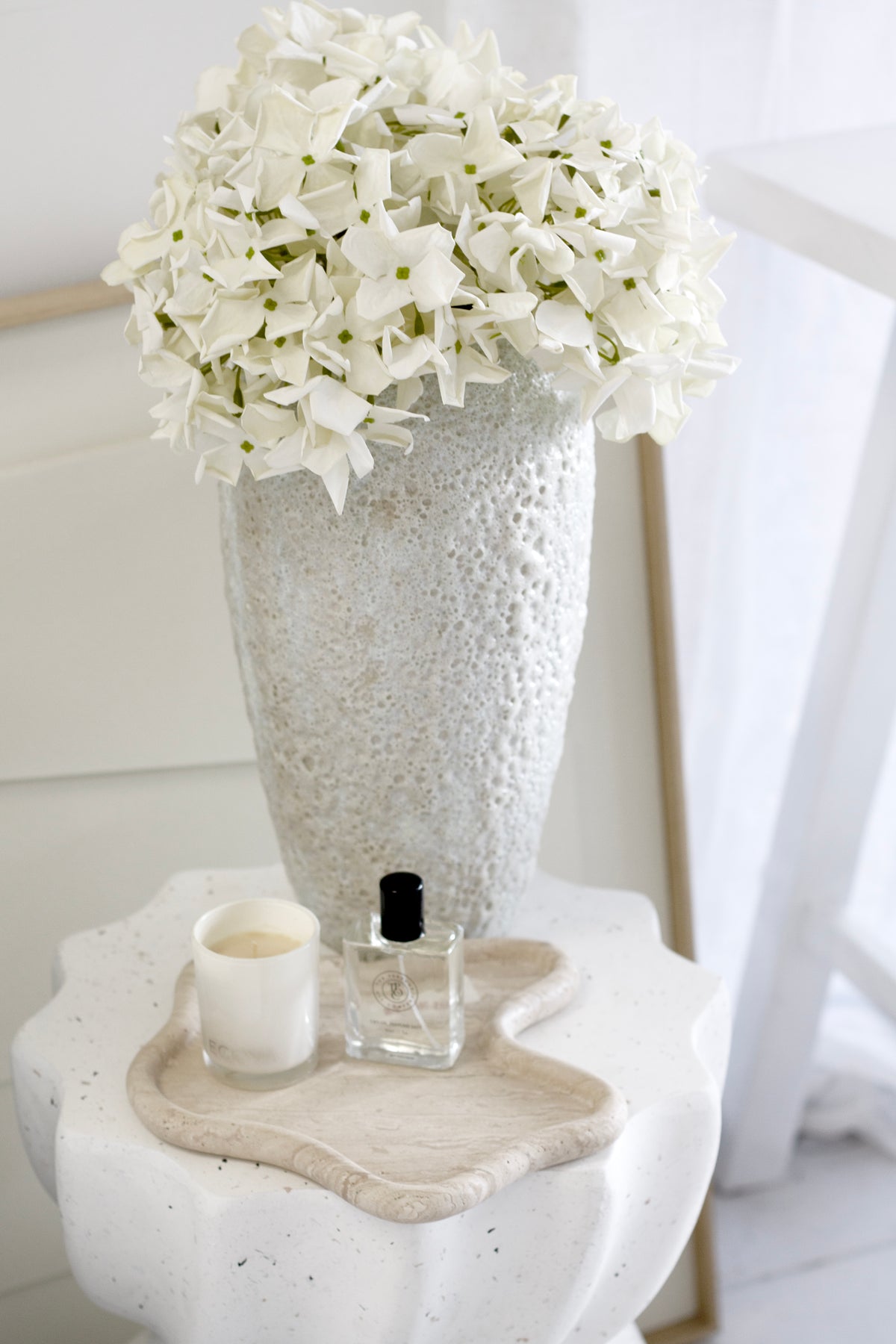 White flowers in a textured vase on a white surface with a candle and small bottle.