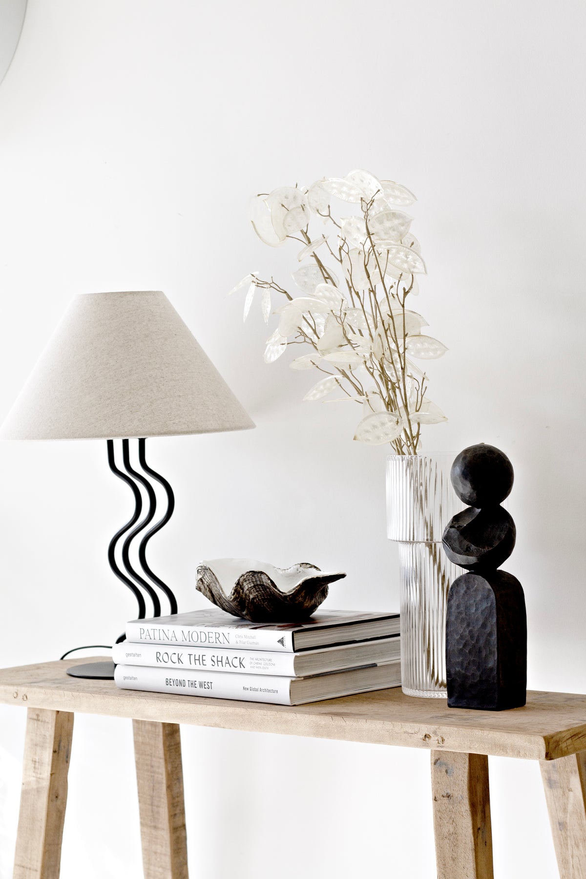 Clam shell dish on stacked books beside a wavy base lamp, dried lunaria stems and a black sculpture
