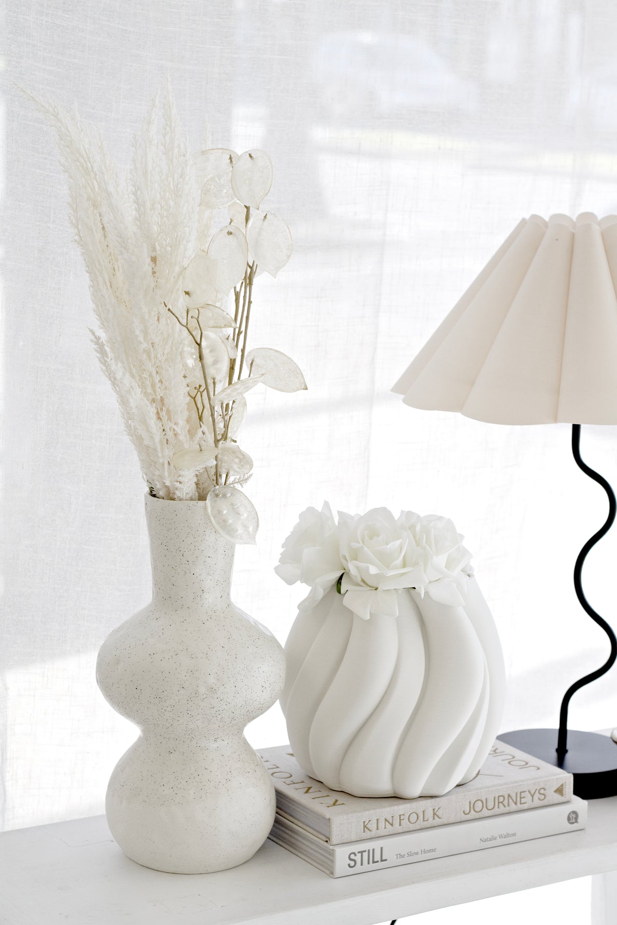 Faux toi toi plumes in a curvy white vase beside a swirl vase and a pleated wavy lamp on a shelf