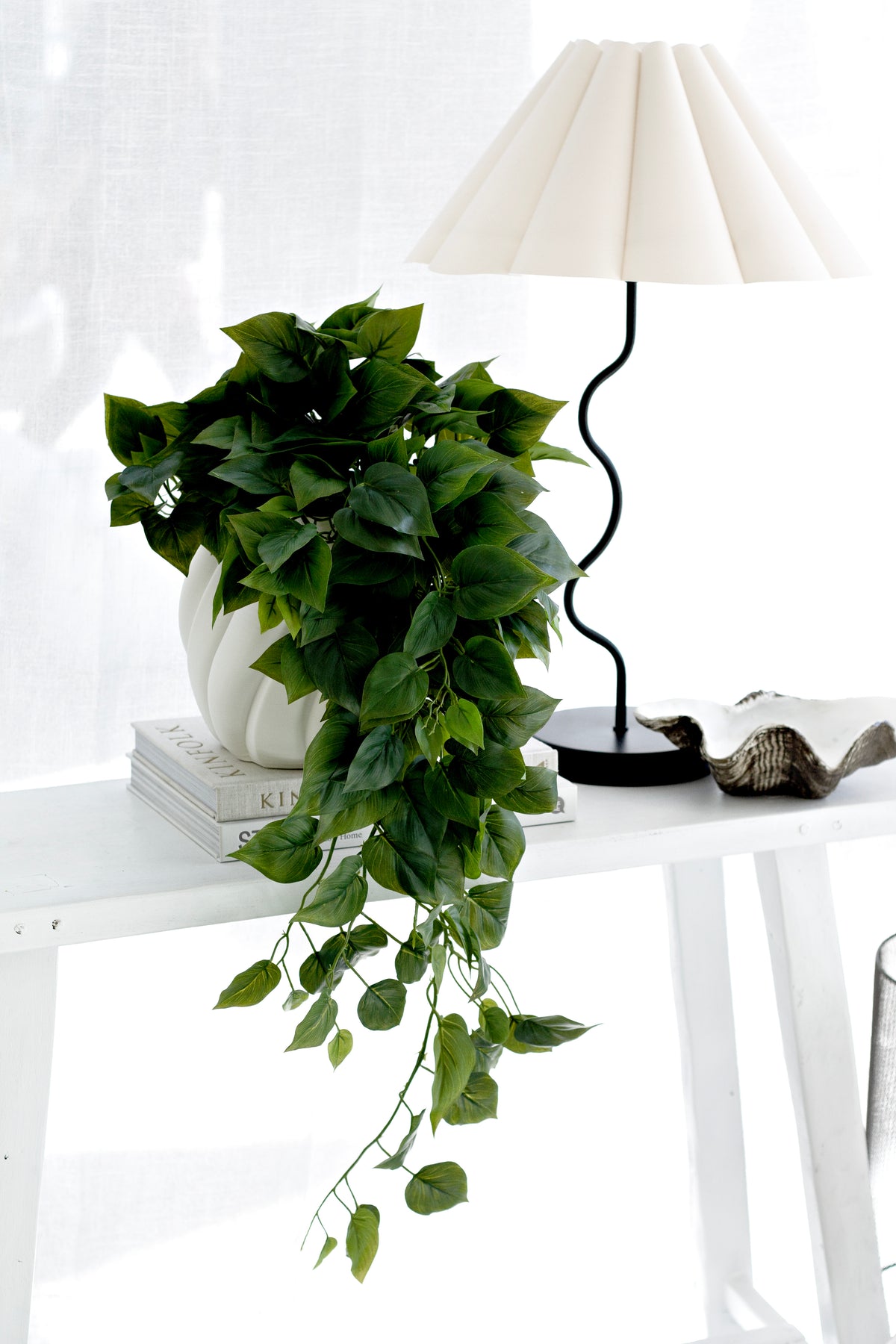 Faux philodendron trailing from a white sculpted pot beside a pleated lamp