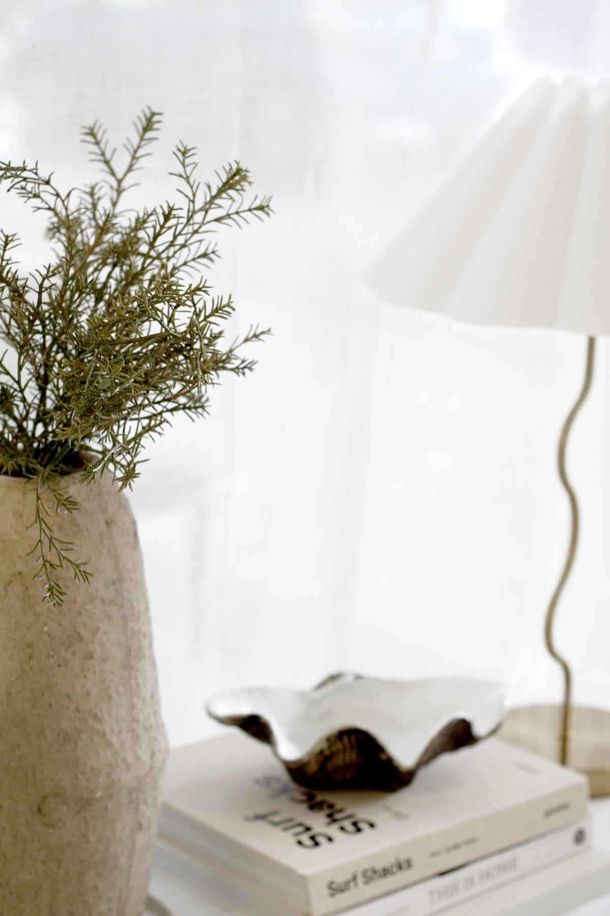 Caru vase of green foliage beside a wavy shell dish and a shaded lamp on books