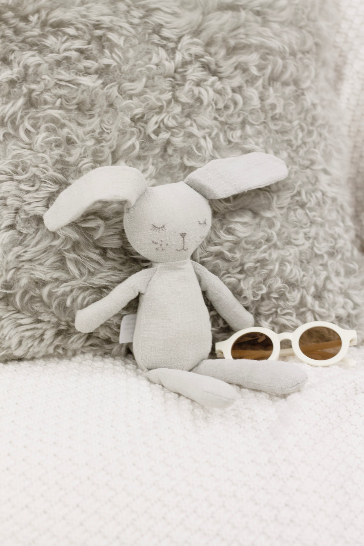 Grey the Bunny soft toy lying on a curly sheepskin rug beside round white sunglasses