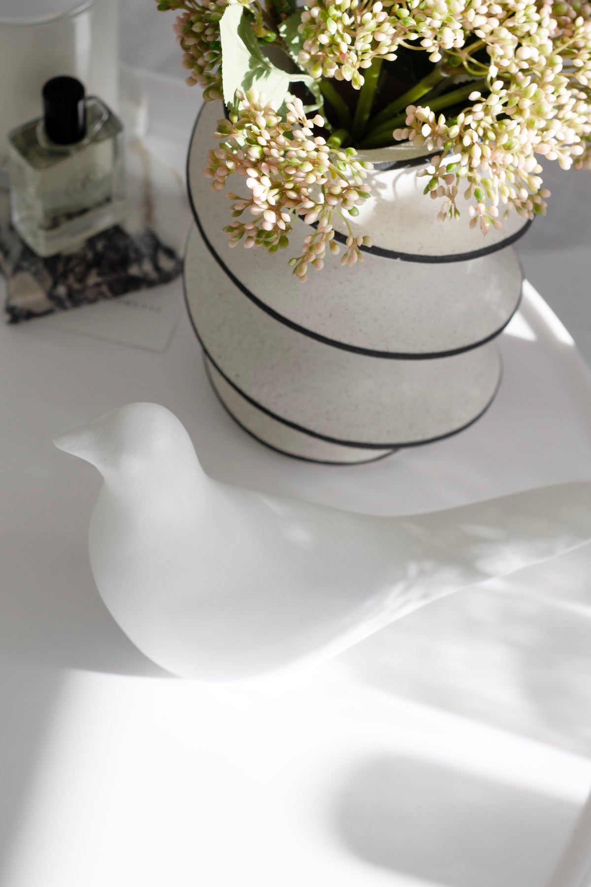 White bird figurine casting a long shadow on a white surface, striped vase of blossom behind