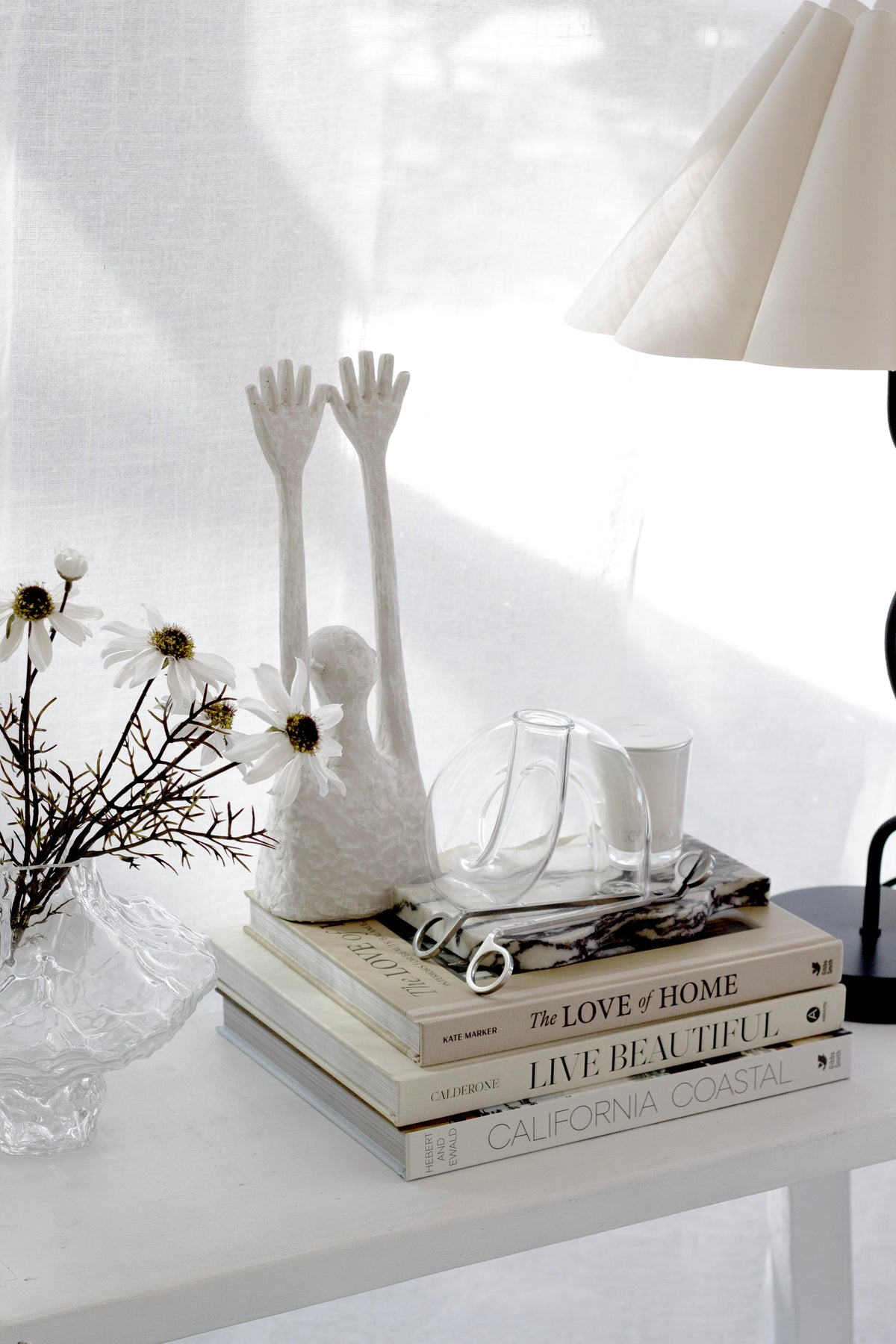 Clear J-Curve vase on a sunlit shelf with a white hand sculpture, dried stems and stacked books