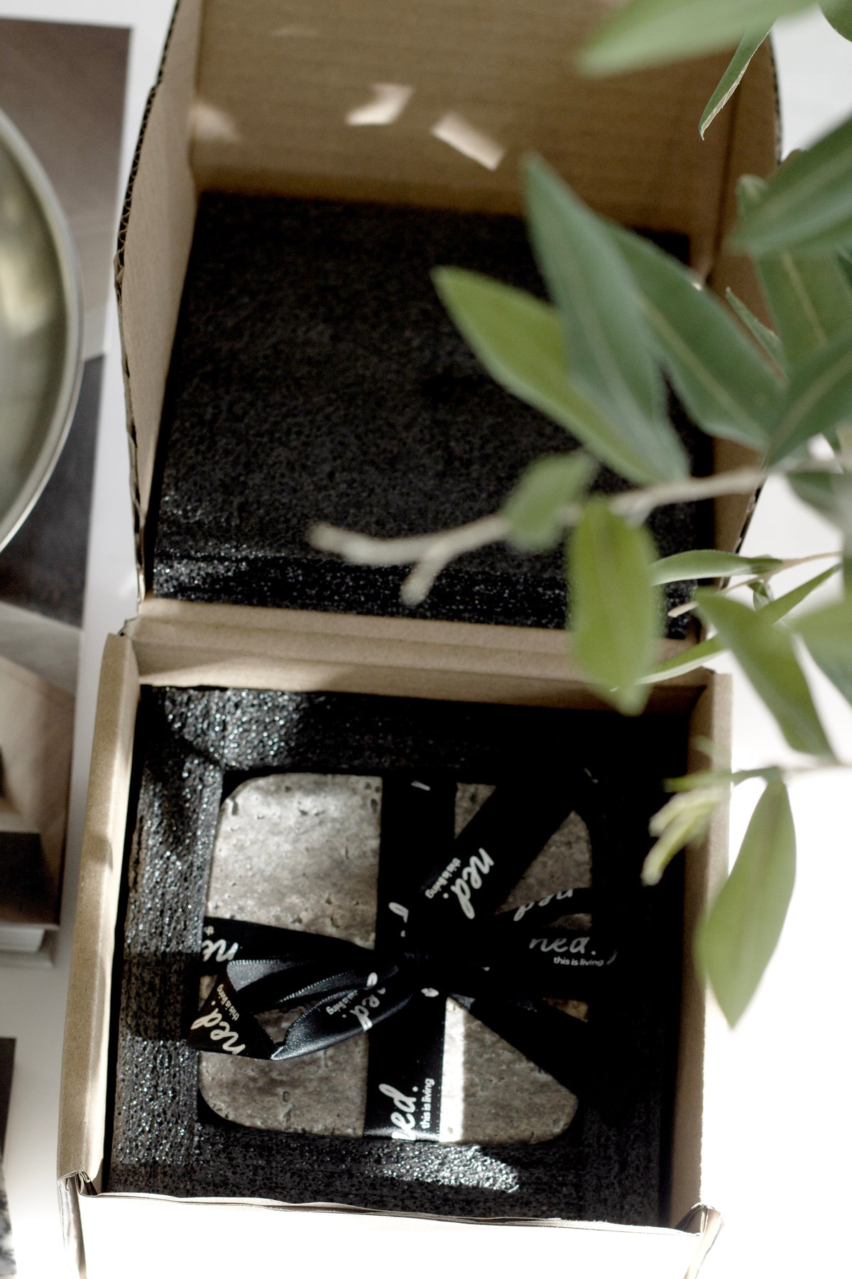 Stone coasters in their open gift box with a black ribbon beside an olive branch