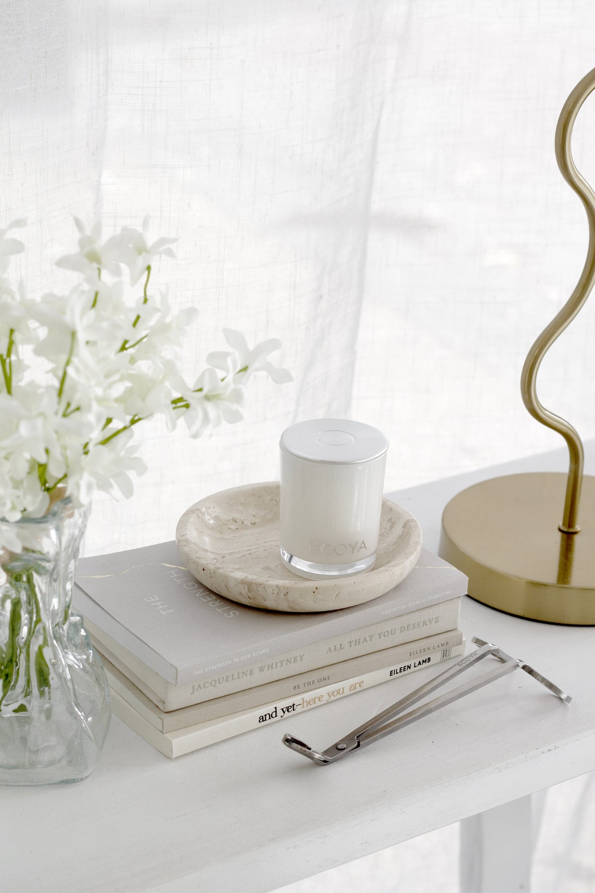 Travertine tray holding a white candle on books, beside a gold lamp and white blossoms