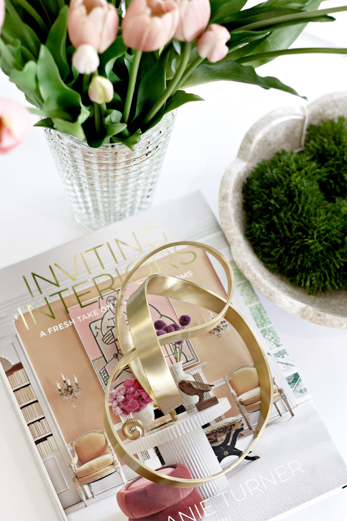 Gold Infinity Loop sculpture on interiors books with pink tulips in a textured vase and a bowl of preserved moss