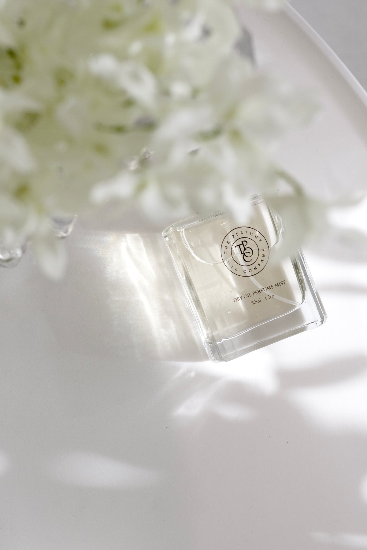 Square glass perfume oil bottle resting on a white surface with blossom and soft shadows