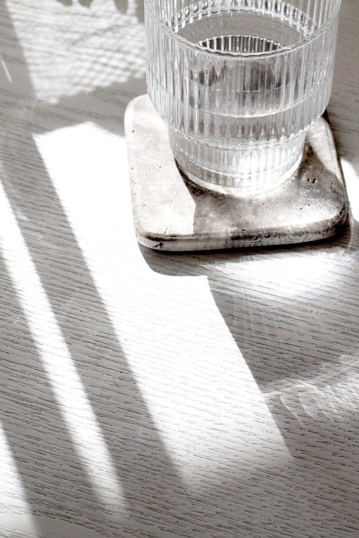 Ribbed glass on a travertine coaster in striped sunlight on timber