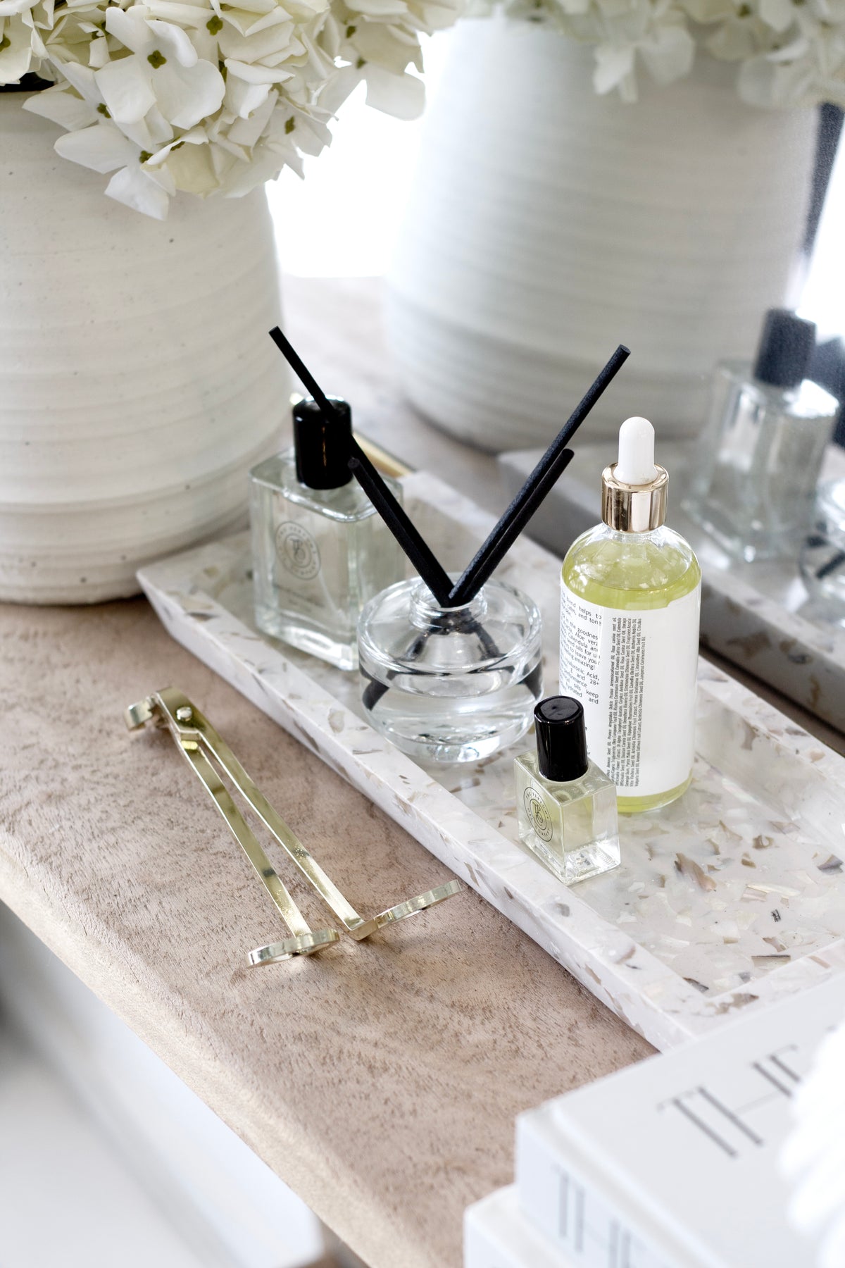 Gold wick trimmer on a stone tray with a reed diffuser, oil bottles and white hydrangeas