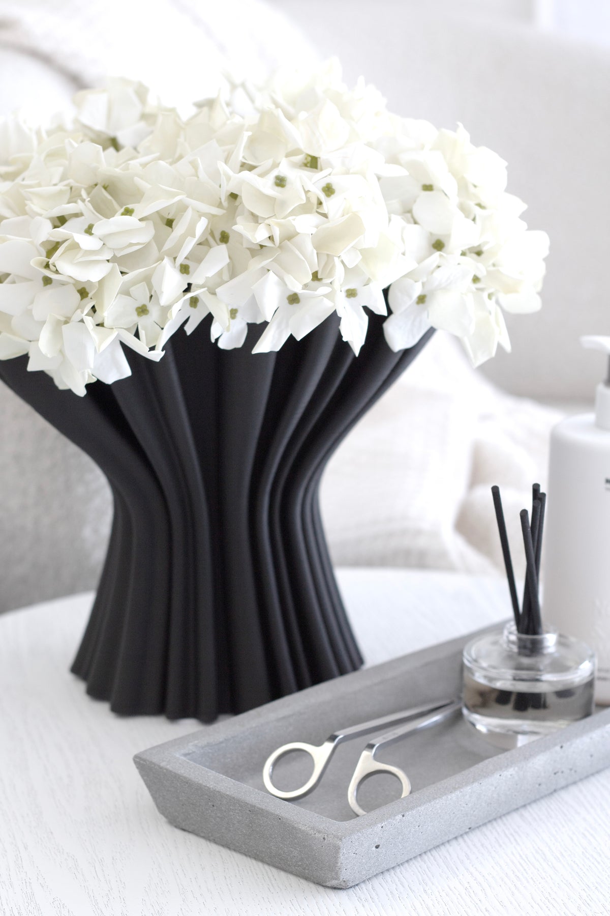 Black Cleo compote vase of white hydrangeas beside a stone tray holding a reed diffuser and scissors