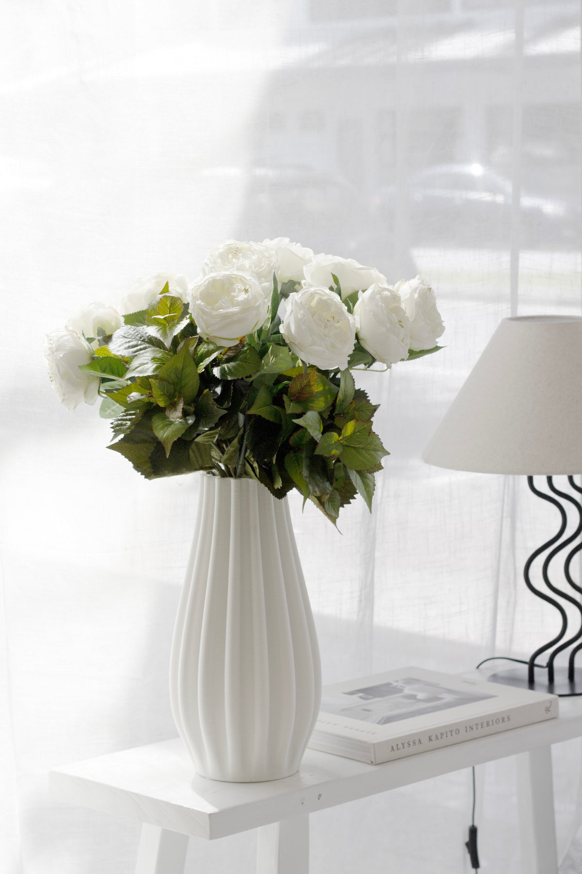 Green leaf bush with white roses in a ribbed vase on a console beside a wavy based lamp