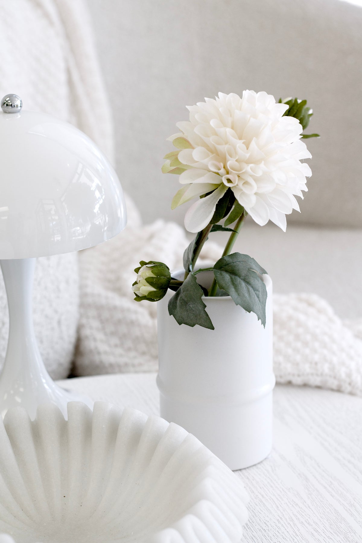 White ceramic bamboo vase holding a white peony beside a scalloped shell dish and a chunky knit throw