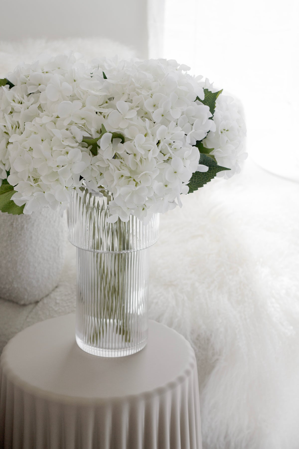 Ribbed glass vase of white hydrangea and roses on a fluted pedestal, faux fur behind