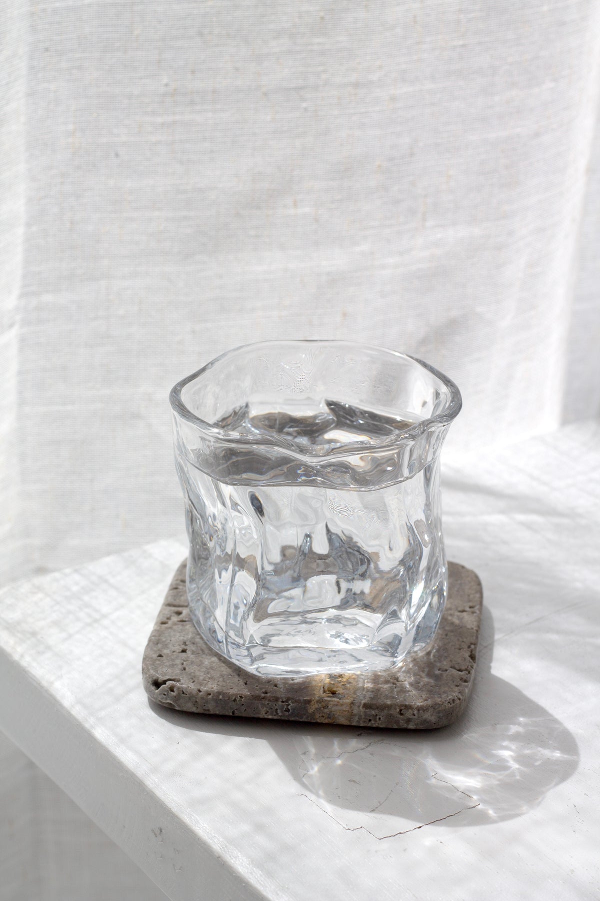 Wavy glass of water on a travertine coaster on white linen