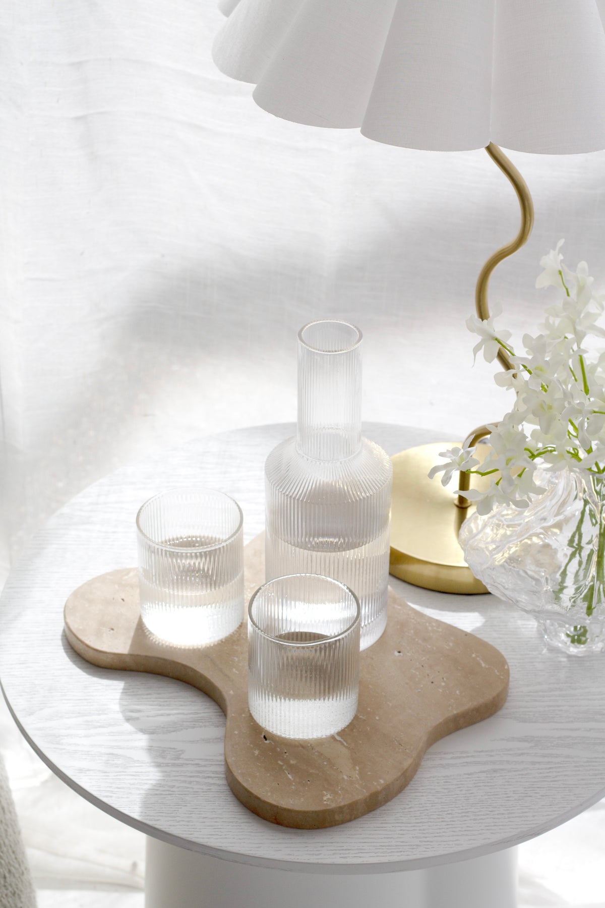 Fluted decanter and tumblers on a travertine tray beneath a scalloped table lamp with a gold base