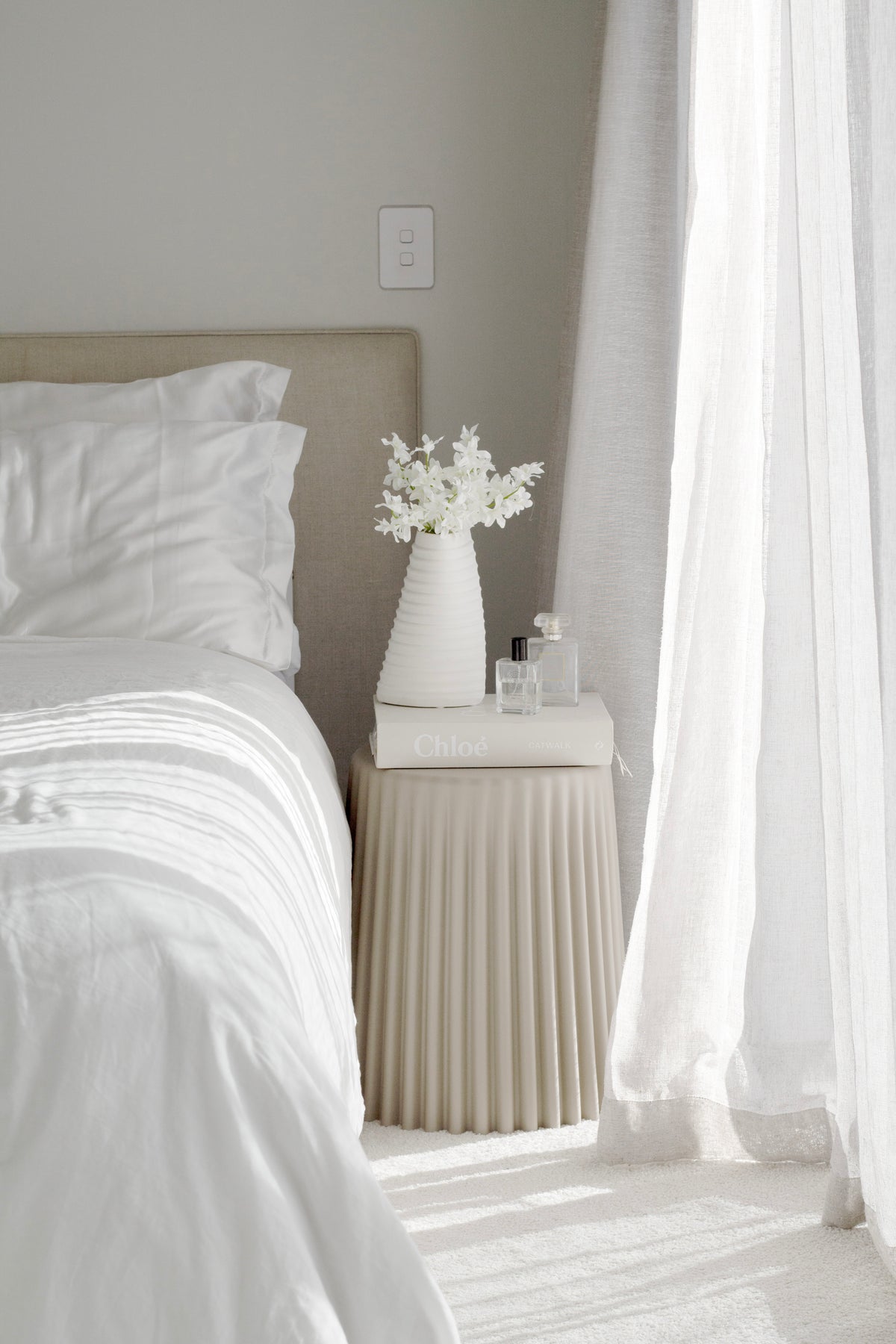 White ribbed stool beside a bed, holding a white vase of flowers against a sheer curtain