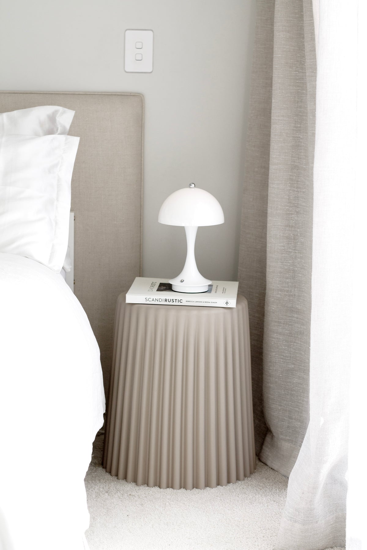 Nude ribbed stool beside an upholstered headboard, a mushroom lamp on a book, curtain behind