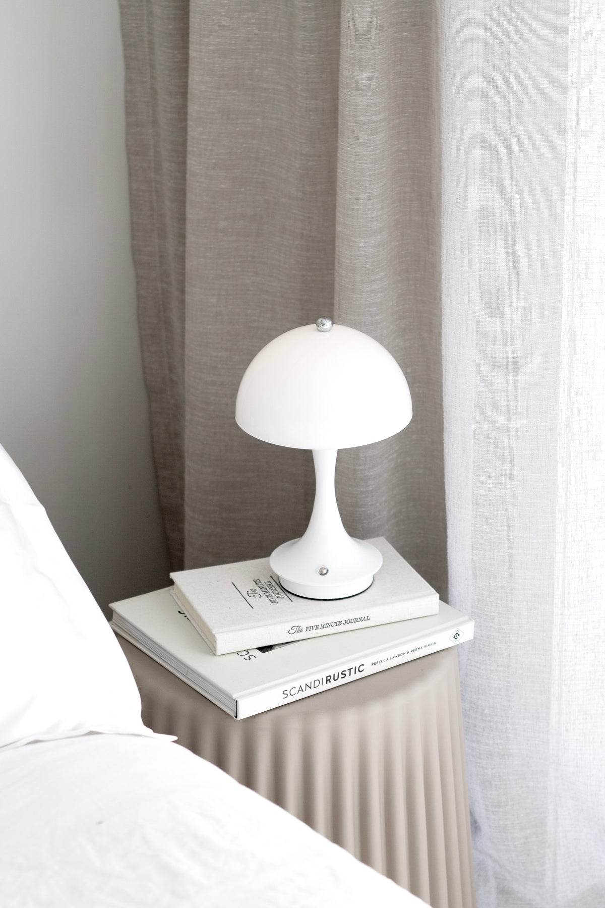 White mushroom lamp lit on books on a fluted bedside table with a sheer curtain behind