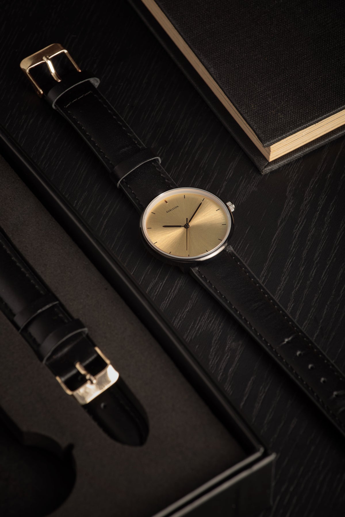 Karlsson Finesse watch on a dark surface beside a spare strap and a black notebook