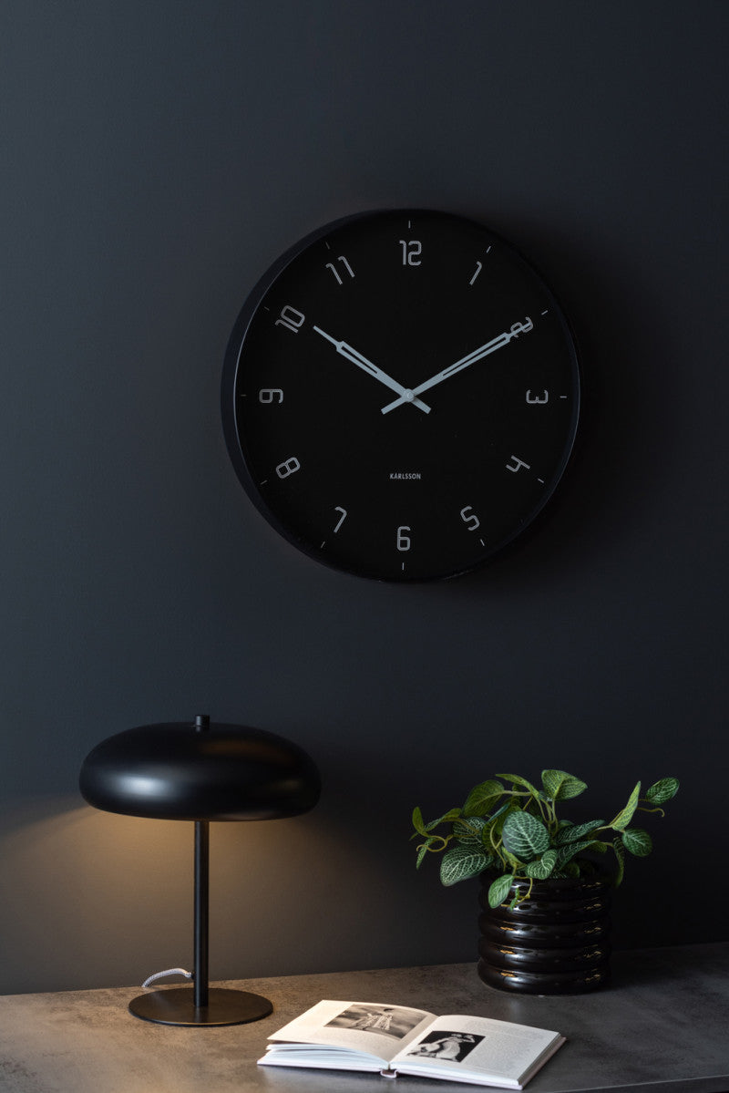 Black Stark wall clock on a dark wall above a desk with a mushroom lamp and a plant