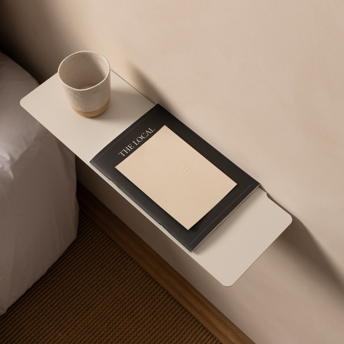 Close-up of a white FOLD Ledge with a cup and notebook beside a bed