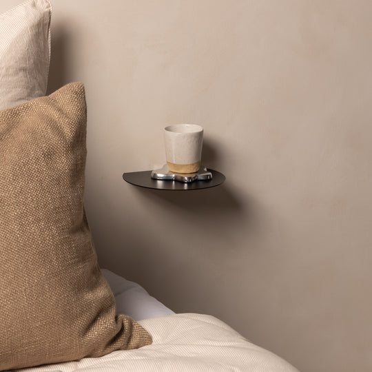 Black FOLD Round Shelf holding a cup beside a bed as a floating bedside shelf