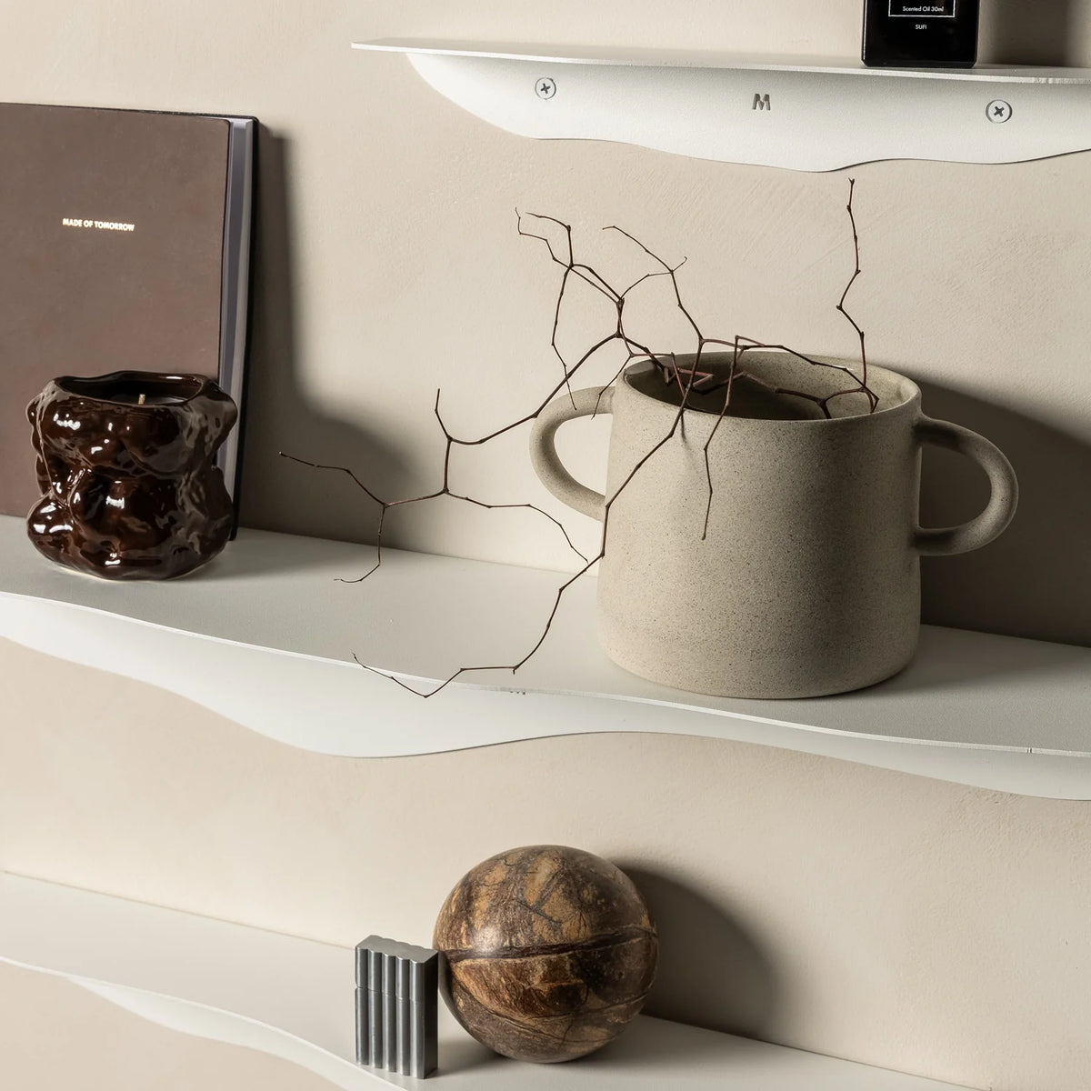 Close view of white wavy shelves holding a book, a glazed vessel, a mug of dried branches and a stone