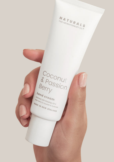 Hand holding the Naturals Coconut & Passion Berry hand cream tube against a warm grey background