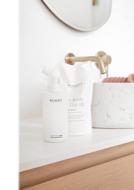 Naturals bottle and refill pouch beside a brass wall tap and textured stone pot on a white vanity