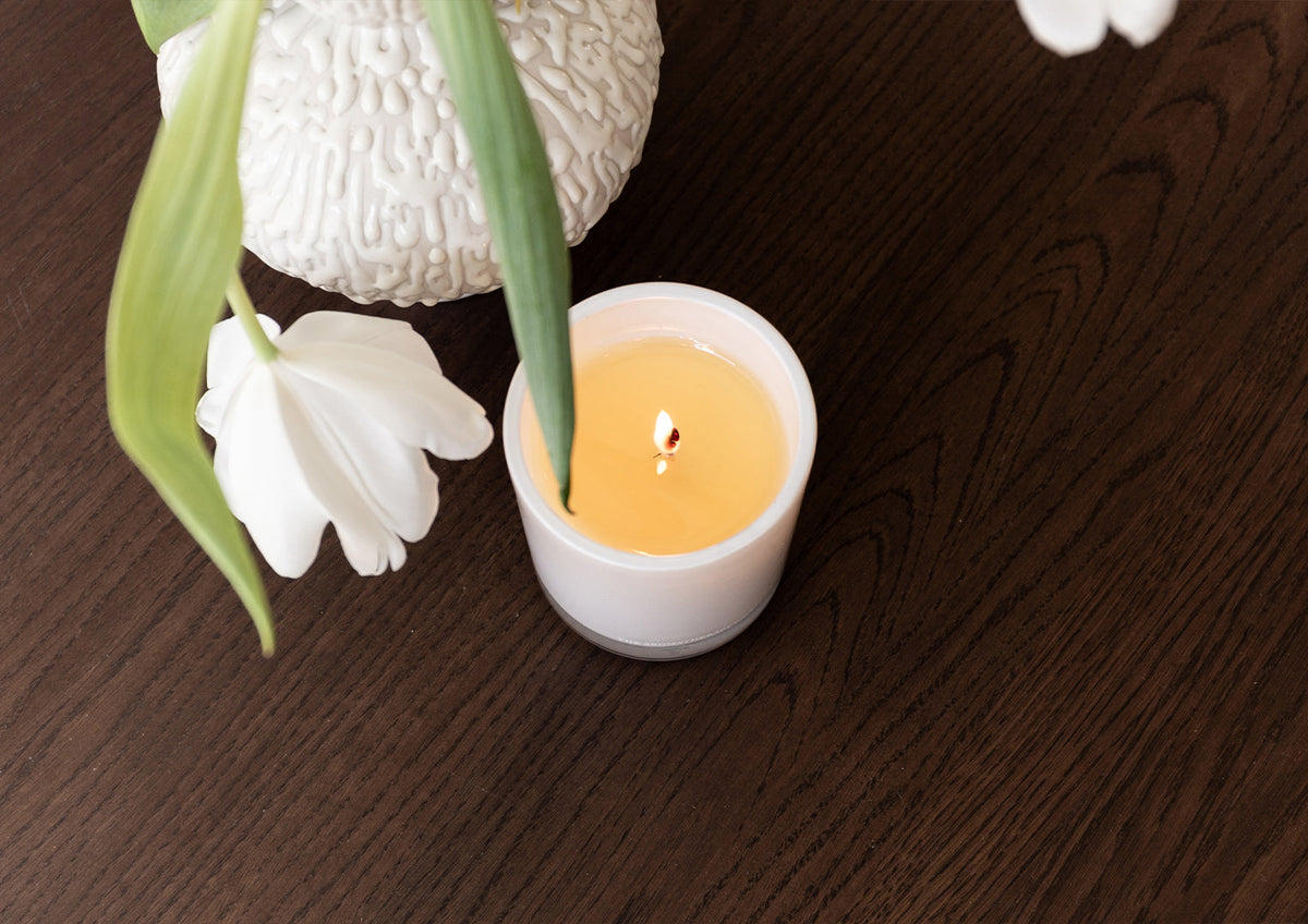 Lit Naturals candle on dark timber beside white tulips and a textured white vase