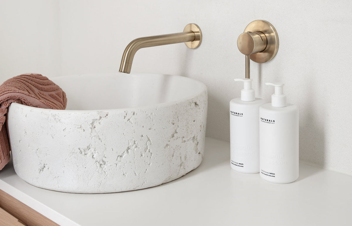 Two Naturals bottles beside a round stone vessel basin under a brushed brass wall spout