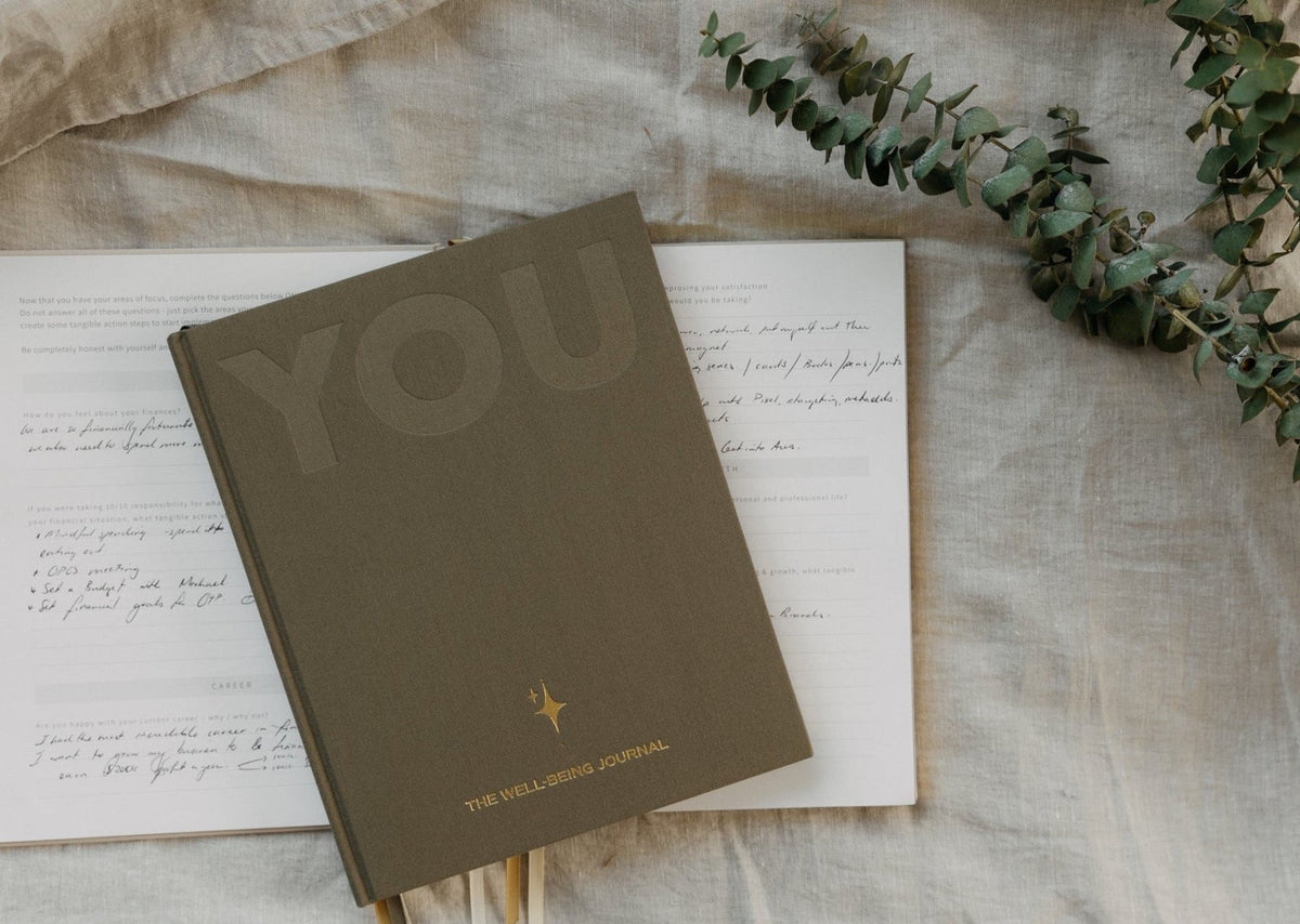 The sand YOU. journal resting on handwritten pages on linen, with eucalyptus stems above