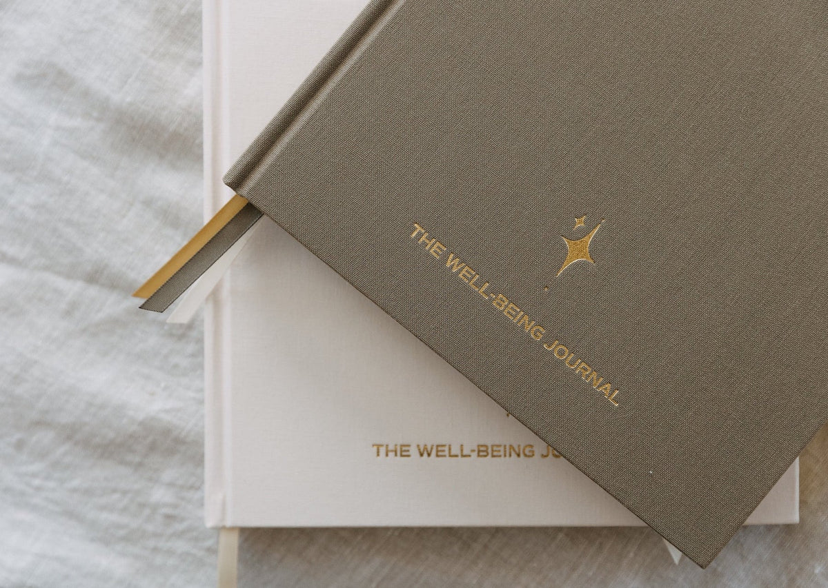 The sand YOU. journal resting on the cream edition, both showing gold foil lettering reading The Well-Being Journal