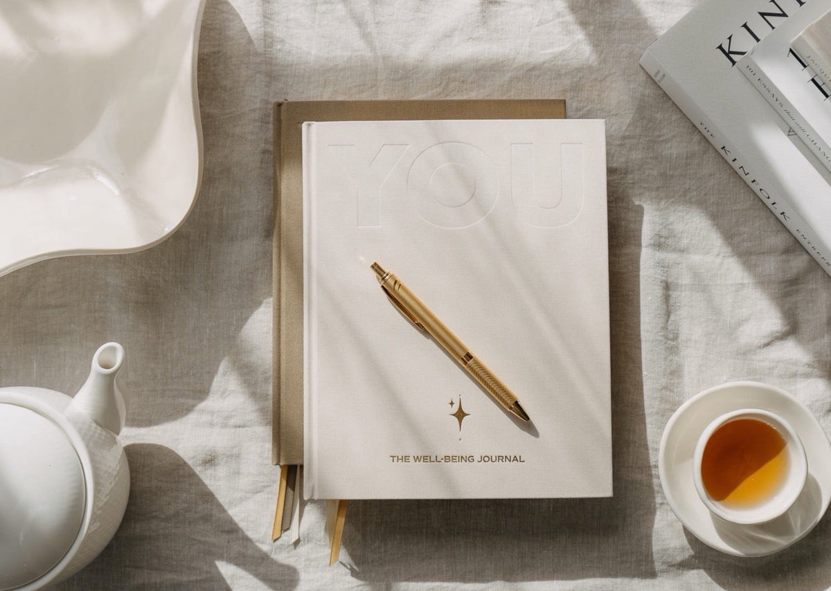 The cream YOU. journal with a gold pen on a linen cloth, styled with a white teapot, a cup of tea and a magazine
