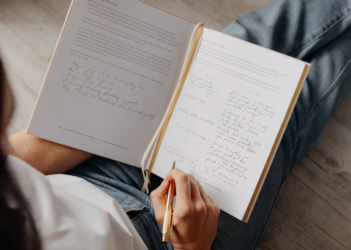 A woman sitting on the floor writing in the YOU. journal resting on her lap