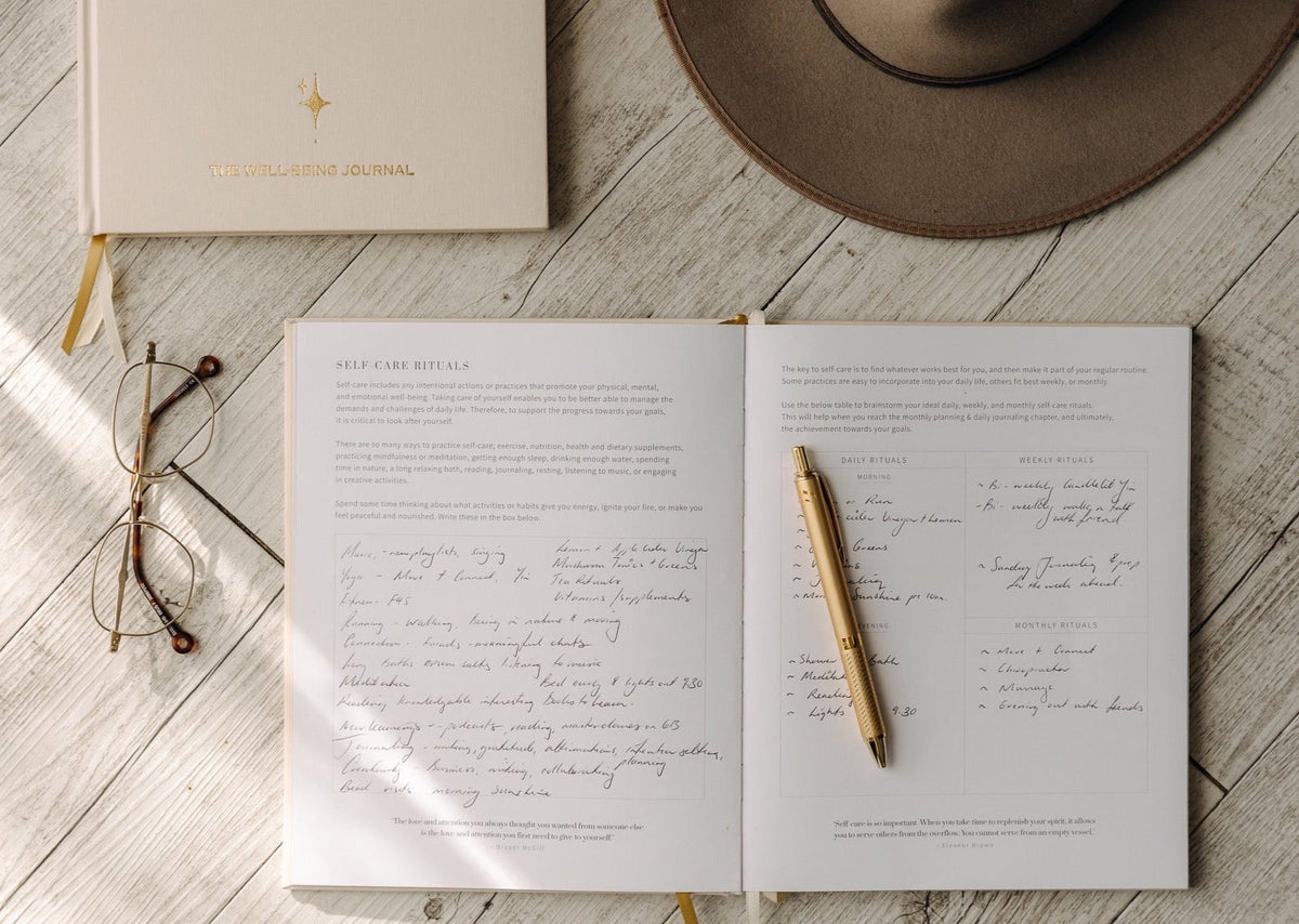 The YOU. journal open on a timber floor with handwritten entries, a gold pen, reading glasses and a felt hat