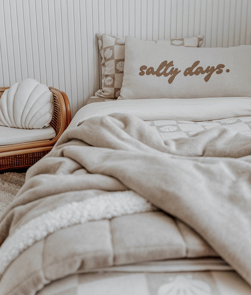 Salty Days cotton pillowcase with stone script lettering on a bed with a shell cushion and knit throw