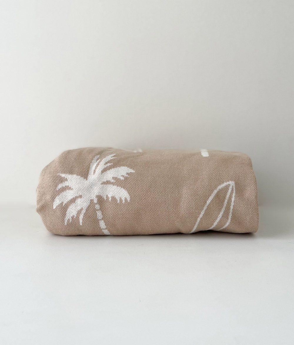 Folded sand Surfing Palm jersey fitted sheet with ivory palm and surfboard print on a white backdrop