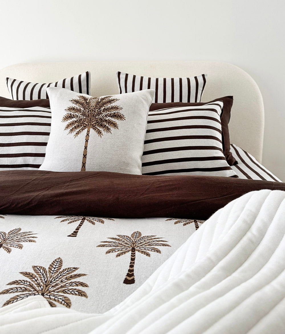 Made bed with chocolate and ivory striped pillowcases behind a palm print cushion