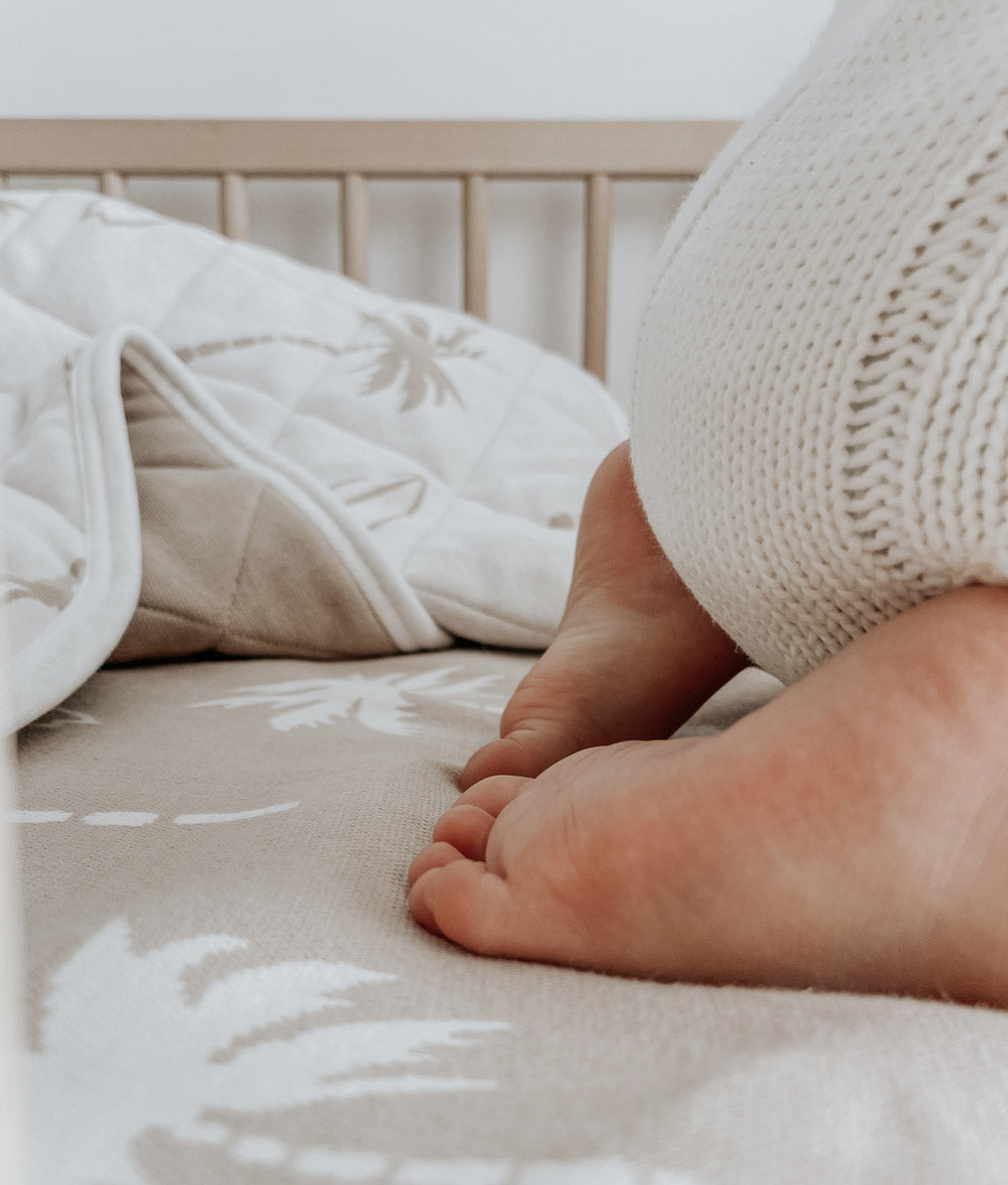 Baby's feet resting on a sand Surfing Palm jersey cot sheet beside a quilted Surfing Palm quilt